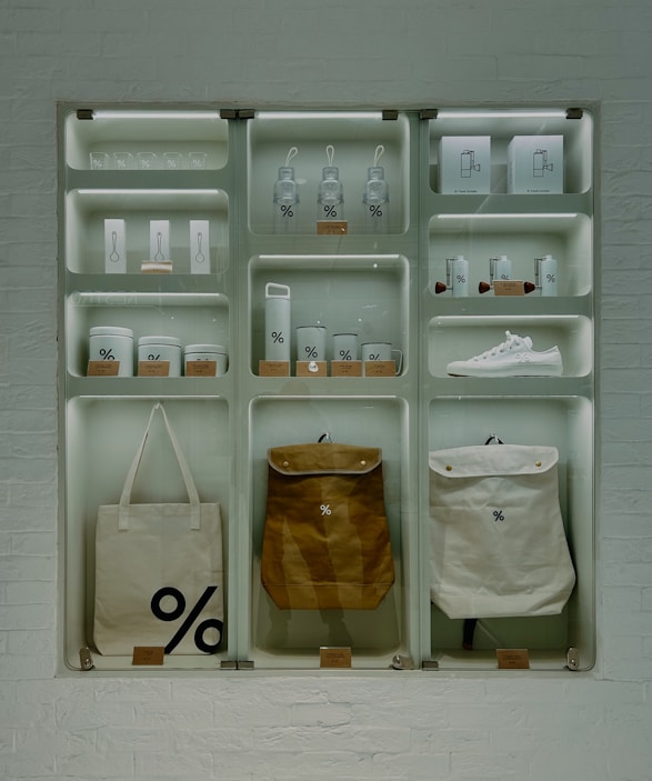 A well-organized display of branded merchandise with a minimalist aesthetic. The items include bottles, cups, a sneaker, bags, and other accessories, all featuring a distinctive percentage symbol on a white or neutral background. The display is set against a white brick wall, giving an industrial feel.