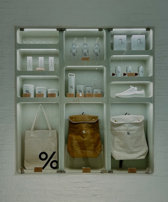 A well-organized display of branded merchandise with a minimalist aesthetic. The items include bottles, cups, a sneaker, bags, and other accessories, all featuring a distinctive percentage symbol on a white or neutral background. The display is set against a white brick wall, giving an industrial feel.