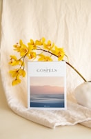 A white book titled 'The Gospels' rests against a soft, beige fabric background. Yellow orchids with reddish centers are elegantly arranged nearby, adding a vibrant touch. The cover of the book features an image of serene, pastel-colored dunes at sunset.