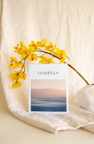 A white book titled 'The Gospels' rests against a soft, beige fabric background. Yellow orchids with reddish centers are elegantly arranged nearby, adding a vibrant touch. The cover of the book features an image of serene, pastel-colored dunes at sunset.