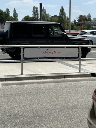 A dark-colored SUV is parked next to a sign that reads 'Hamburg Airport'. The background features other vehicles and a line of tall trees under a partly cloudy sky.
