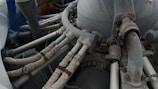 A detailed view of a mechanical engine component with several pipes and tubes. The metal surfaces show signs of wear and rust, while the tubes are wrapped in a protective material. Various bolts and connectors are visible, indicating a complex machinery setup.