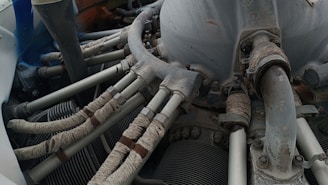 A detailed view of a mechanical engine component with several pipes and tubes. The metal surfaces show signs of wear and rust, while the tubes are wrapped in a protective material. Various bolts and connectors are visible, indicating a complex machinery setup.