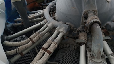 A detailed view of a mechanical engine component with several pipes and tubes. The metal surfaces show signs of wear and rust, while the tubes are wrapped in a protective material. Various bolts and connectors are visible, indicating a complex machinery setup.