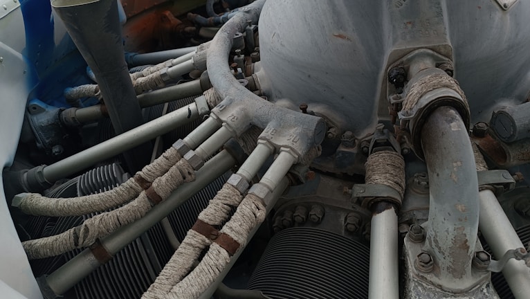 A detailed view of a mechanical engine component with several pipes and tubes. The metal surfaces show signs of wear and rust, while the tubes are wrapped in a protective material. Various bolts and connectors are visible, indicating a complex machinery setup.