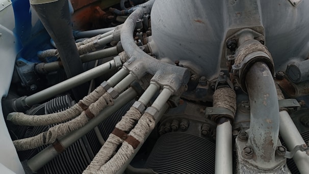 A detailed view of a mechanical engine component with several pipes and tubes. The metal surfaces show signs of wear and rust, while the tubes are wrapped in a protective material. Various bolts and connectors are visible, indicating a complex machinery setup.