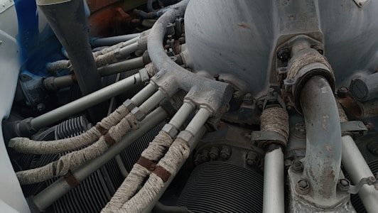 A detailed view of a mechanical engine component with several pipes and tubes. The metal surfaces show signs of wear and rust, while the tubes are wrapped in a protective material. Various bolts and connectors are visible, indicating a complex machinery setup.