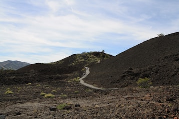A winding path cuts through a rugged volcanic landscape with dark, rocky hills. Sparse patches of greenery are scattered across the terrain, and a few people are visible walking along the path in the distance. The sky is mostly clear with soft clouds, providing a contrast to the dark earth.