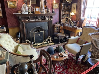 A Victorian-style room featuring a fireplace adorned with various ornaments such as candlesticks and small sculptures. The room is filled with antique furniture including a pair of upholstered armchairs and a small wooden table with hats and a magnifying glass on it. A violin rests on sheet music atop a wooden desk next to shelves filled with glass bottles and decorations. The room’s rich decor includes paintings, an ornate rug, and elaborate wallpaper, contributing to an old-world charm.