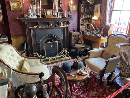 A Victorian-style room featuring a fireplace adorned with various ornaments such as candlesticks and small sculptures. The room is filled with antique furniture including a pair of upholstered armchairs and a small wooden table with hats and a magnifying glass on it. A violin rests on sheet music atop a wooden desk next to shelves filled with glass bottles and decorations. The room’s rich decor includes paintings, an ornate rug, and elaborate wallpaper, contributing to an old-world charm.