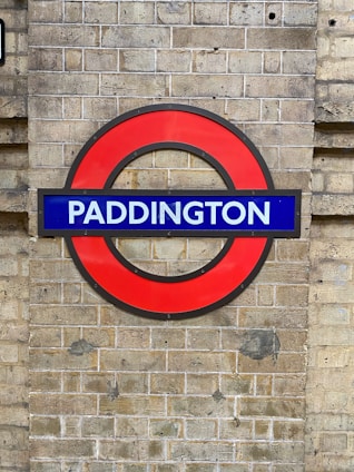 A brick wall features a prominent circular red sign with a blue rectangular banner across the center. The word 'Paddington' is written in bold white letters on the blue section.