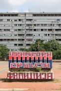 A large, multi-story apartment building with numerous windows and air conditioning units. In the foreground, there's a brick structure painted with the colors red, blue, and white, featuring bold Cyrillic text. The scene includes some greenery and a cloudy sky.