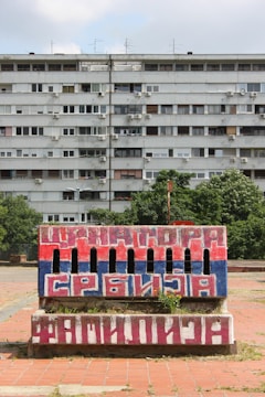 A large, multi-story apartment building with numerous windows and air conditioning units. In the foreground, there's a brick structure painted with the colors red, blue, and white, featuring bold Cyrillic text. The scene includes some greenery and a cloudy sky.
