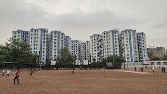 A wide view of a residential complex with multiple tall buildings in a uniform style, predominantly white with blue accents. In the foreground, an open sandy area is visible where people are playing sports, possibly cricket, as some individuals hold bats. The ground is surrounded by a line of trees. The sky is overcast, creating a muted atmosphere.