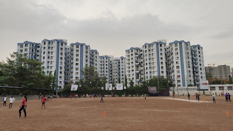 A wide view of a residential complex with multiple tall buildings in a uniform style, predominantly white with blue accents. In the foreground, an open sandy area is visible where people are playing sports, possibly cricket, as some individuals hold bats. The ground is surrounded by a line of trees. The sky is overcast, creating a muted atmosphere.