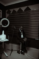 A person dressed in a suit is sitting on a stool in a room with acoustic panels and sound equipment. The lighting is dim, creating a moody atmosphere. A microphone on a stand and a glass on a table are visible, suggesting a recording or music studio setting.