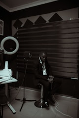 A person dressed in a suit is sitting on a stool in a room with acoustic panels and sound equipment. The lighting is dim, creating a moody atmosphere. A microphone on a stand and a glass on a table are visible, suggesting a recording or music studio setting.