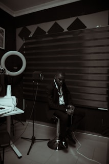 A person dressed in a suit is sitting on a stool in a room with acoustic panels and sound equipment. The lighting is dim, creating a moody atmosphere. A microphone on a stand and a glass on a table are visible, suggesting a recording or music studio setting.
