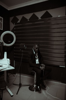 A person dressed in a suit is sitting on a stool in a room with acoustic panels and sound equipment. The lighting is dim, creating a moody atmosphere. A microphone on a stand and a glass on a table are visible, suggesting a recording or music studio setting.