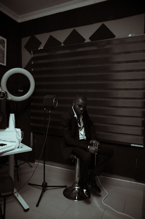 A person dressed in a suit is sitting on a stool in a room with acoustic panels and sound equipment. The lighting is dim, creating a moody atmosphere. A microphone on a stand and a glass on a table are visible, suggesting a recording or music studio setting.