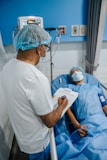 A healthcare professional is standing beside a patient who is lying in a hospital bed. Medical equipment is visible in the background.