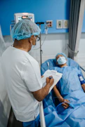 A healthcare professional is standing beside a patient who is lying in a hospital bed. The professional is wearing a surgical cap, mask, and white coat, while holding a clipboard. The patient is covered with a blue sheet and also wears a mask. Medical equipment is visible in the background.