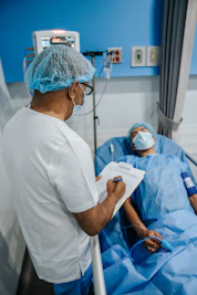 A healthcare professional is standing beside a patient who is lying in a hospital bed. The professional is wearing a surgical cap, mask, and white coat, while holding a clipboard. The patient is covered with a blue sheet and also wears a mask. Medical equipment is visible in the background.