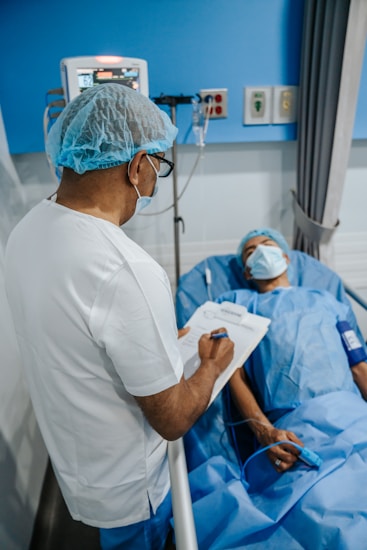 A healthcare professional is standing beside a patient who is lying in a hospital bed. The professional is wearing a surgical cap, mask, and white coat, while holding a clipboard. The patient is covered with a blue sheet and also wears a mask. Medical equipment is visible in the background.