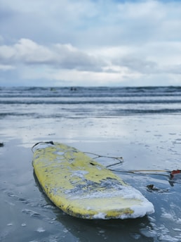 A weathered yellow surfboard lies on the wet sand at the edge of the sea, with patches of foam attached. In the background, the ocean waves are rolling under a cloudy sky, creating a calm yet slightly overcast atmosphere.
