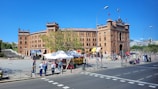 A grand, historic bullring with intricate architectural details, featuring a brick facade and multiple towers. People are walking around the spacious plaza in front of the building, and a few market stalls with white canopies are set up, offering goods. A large tree provides some greenery to the area.