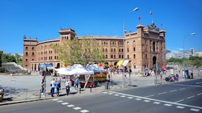 A grand, historic bullring with intricate architectural details, featuring a brick facade and multiple towers. People are walking around the spacious plaza in front of the building, and a few market stalls with white canopies are set up, offering goods. A large tree provides some greenery to the area.