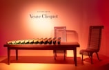 A collection of champagne bottles arranged in a neat line on a wooden table. The setting includes a unique wooden chair design, with the backdrop illuminated by a warm orange-pink gradient. The brand name 'Veuve Clicquot' is prominently displayed on the wall above the table.