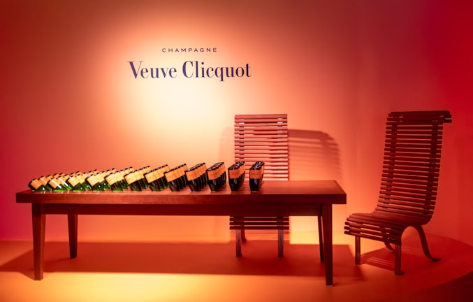 A collection of champagne bottles arranged in a neat line on a wooden table. The setting includes a unique wooden chair design, with the backdrop illuminated by a warm orange-pink gradient. The brand name 'Veuve Clicquot' is prominently displayed on the wall above the table.