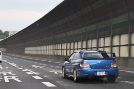 A blue sports car is driving on a highway with a tunnel-like noise barrier. The road markings include Japanese characters, indicating the location might be in Japan. The car's distinct spoiler and sleek design suggest it is designed for performance. Surrounding the road are urban elements and some vegetation in the distance.