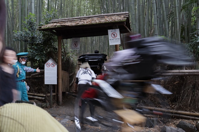 A bamboo forest serves as the backdrop with signs indicating restricted access. In the foreground, a security officer wearing a blue uniform and mask is gesturing towards the signs. A blurred figure on a bicycle is passing quickly, creating a sense of motion.