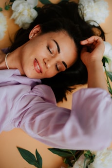 A woman with dark hair and closed eyes appears relaxed, lying on a soft surface surrounded by white peonies. She is wearing a light purple blouse and a pearl necklace, with a gentle smile on her face.