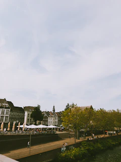 A riverside setting features a row of historic buildings with detailed architecture. A tree-lined promenade with people walking and sitting is visible alongside a waterway. Outdoor seating includes white umbrellas, suggesting a café or dining area.