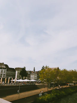 A riverside setting features a row of historic buildings with detailed architecture. A tree-lined promenade with people walking and sitting is visible alongside a waterway. Outdoor seating includes white umbrellas, suggesting a café or dining area.