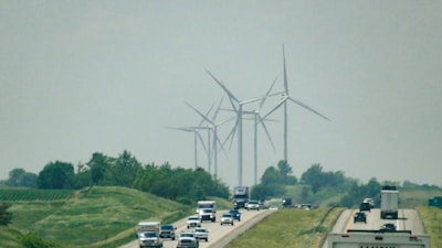 A highway with multiple vehicles, including trucks and cars, surrounded by green fields and trees. Several large wind turbines are visible in the background, blending into the overcast sky.