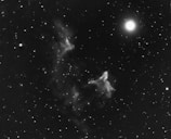 An intricate interstellar scene featuring a bright star illuminating the right side, surrounded by various other smaller stars scattered across the dark cosmic expanse. Wisps of nebulae appear to drift and swirl in the center, creating ghostly shapes against the vastness of space.
