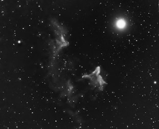 An intricate interstellar scene featuring a bright star illuminating the right side, surrounded by various other smaller stars scattered across the dark cosmic expanse. Wisps of nebulae appear to drift and swirl in the center, creating ghostly shapes against the vastness of space.