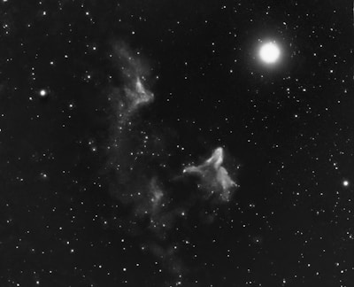 An intricate interstellar scene featuring a bright star illuminating the right side, surrounded by various other smaller stars scattered across the dark cosmic expanse. Wisps of nebulae appear to drift and swirl in the center, creating ghostly shapes against the vastness of space.