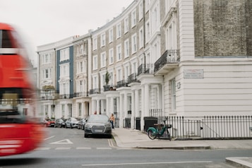 A row of elegant townhouses lines a city street, with classic architectural features such as columns and wrought iron balconies. A red double-decker bus is seen blurring by in motion on the street, while cars are parked along the sidewalk. A bicycle is parked by the railing, and a person walks along the sidewalk. The street sign reads Kensington Park Road W11.