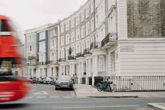 A row of elegant townhouses lines a city street, with classic architectural features such as columns and wrought iron balconies. A red double-decker bus is seen blurring by in motion on the street, while cars are parked along the sidewalk. A bicycle is parked by the railing, and a person walks along the sidewalk. The street sign reads Kensington Park Road W11.