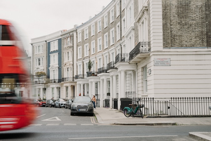 A row of elegant townhouses lines a city street, with classic architectural features such as columns and wrought iron balconies. A red double-decker bus is seen blurring by in motion on the street, while cars are parked along the sidewalk. A bicycle is parked by the railing, and a person walks along the sidewalk. The street sign reads Kensington Park Road W11.