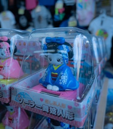 Several figurines are displayed in clear plastic boxes, featuring dolls dressed in traditional attire with vivid colors and intricate designs. The dolls have wide eyes and are wearing blue and pink clothing with floral patterns. They appear to be on a store shelf with various products in the background, slightly out of focus.