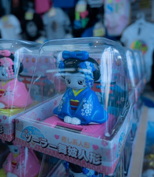 Several figurines are displayed in clear plastic boxes, featuring dolls dressed in traditional attire with vivid colors and intricate designs. The dolls have wide eyes and are wearing blue and pink clothing with floral patterns. They appear to be on a store shelf with various products in the background, slightly out of focus.