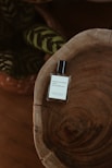 A minimalist and elegant perfume bottle sits atop a rustic wooden surface with visible grain patterns. In the background, there are dark green leaves with lighter stripes, adding a touch of nature to the composition.