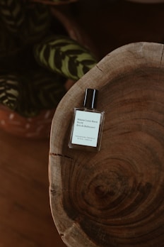 A minimalist and elegant perfume bottle sits atop a rustic wooden surface with visible grain patterns. In the background, there are dark green leaves with lighter stripes, adding a touch of nature to the composition.