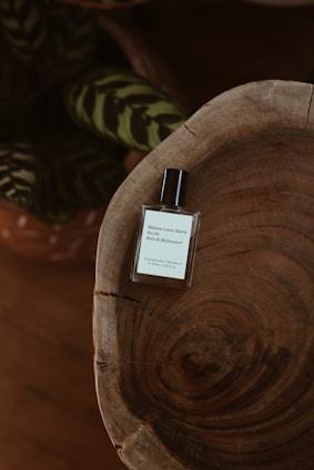 A minimalist and elegant perfume bottle sits atop a rustic wooden surface with visible grain patterns. In the background, there are dark green leaves with lighter stripes, adding a touch of nature to the composition.
